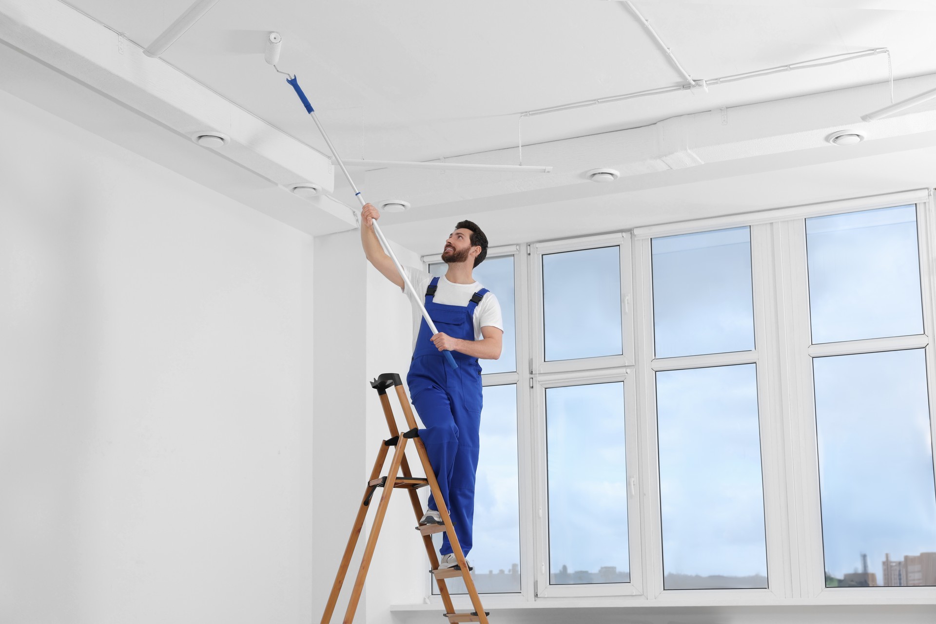 Handyman painting ceiling with roller on step ladder in room